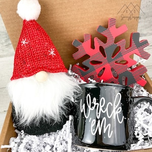 May include: A Christmas gift box filled with white shredded paper. The box contains a black mug with white lettering that says "wreck 'em", a red and black plaid snowflake, and a red and white gnome.