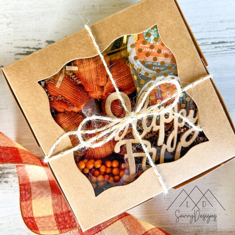 May include: A square brown cardboard gift box with a leaf-shaped window tied with twine. Inside is a fabric pumpkin, a Starbucks Pumpkin Spice coffee pod, and a wooden cutout with the text Happy Fall, all resting on brown paper filler.