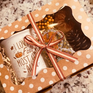 May include: A rectangular gift box with a brown and white polka dot pattern and a clear window, tied with a striped ribbon. Inside is a white mug with the text I LOVE FALL MOST OF ALL and a pumpkin graphic, alongside a pinecone and decorative berries.