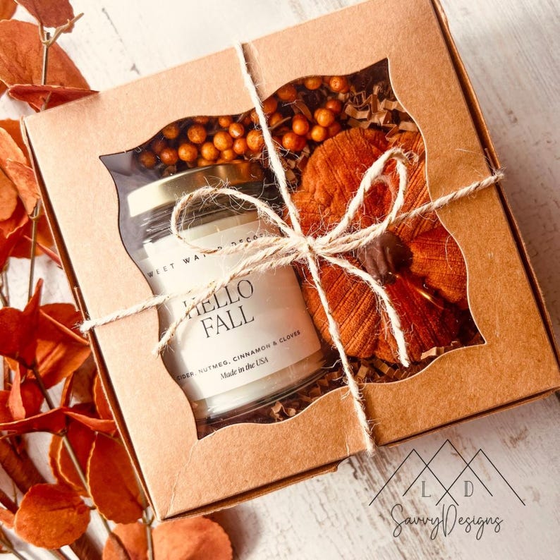 May include: A square cardboard gift box with a window containing a glass candle jar labeled HELLO FALL with notes of nutmeg, cinnamon, and cloves, alongside a small orange fabric pumpkin and decorative berries, tied with twine.