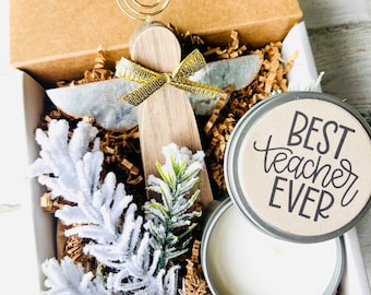 May include: A gift box filled with a small wooden angel figurine with a gold bow, a white candle in a tin, and a tin with the text "Best Teacher Ever" on it. The box is filled with brown shredded paper and white and green sprigs.