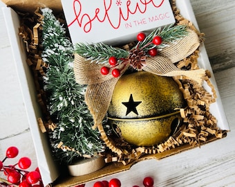 May include: A gift box filled with Christmas decorations, including a small artificial pine tree, a gold bell with a star, and a sign that reads "Believe in the Magic".