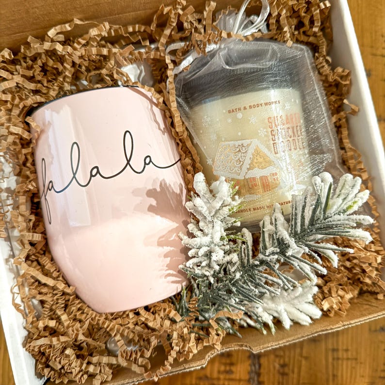 Op de afbeelding: Een roze mok met het woord "falala" in zwarte cursieve letters, een kleine witte kaars met een gingerbread house-ontwerp en de tekst "Sugar Cookie Dough" op het etiket, en een takje met bevroren dennen in een cadeaudoos gevuld met bruin verpakkingspapier.