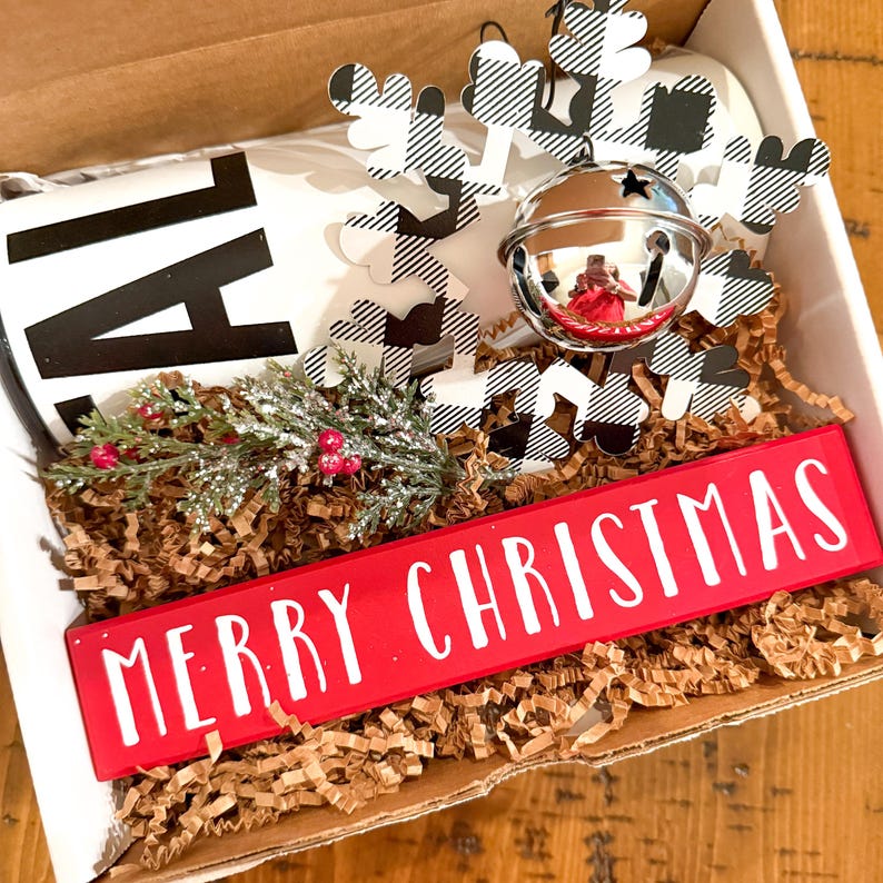 Op de afbeelding: Een witte mok met zwarte letters die "AL" zeggen, een rood houten bord met de tekst "Merry Christmas", een zwart-wit geruit sneeuwvlok ornament met een zilveren bel en bruin kreukpapier.