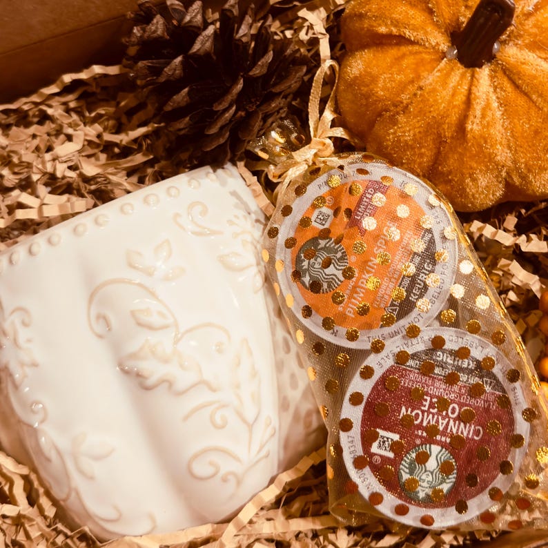 May include: A white ceramic mug with a raised pumpkin-shaped texture, a pinecone, a small orange velvet pumpkin, and a gold-dotted organza bag containing two Starbucks coffee pods, one labeled Pumpkin Spice and one labeled Cinnamon Dolce.