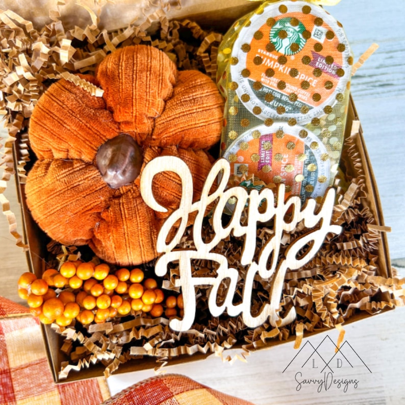 May include: A gift box containing a plush orange corduroy pumpkin, two Starbucks Pumpkin Spice coffee pods in a gold polka-dot mesh bag, a cluster of small orange berries, and a wooden cutout sign with the script text Happy Fall.