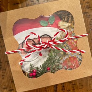 Op de afbeelding: Een bruine kartonnen doos met een transparant venster dat een kerstman-uitsparing, groene takjes, rode bessen en een rode en witte touw strik laat zien. De doos bevat ook een klein pakketje Starbucks-koffie.