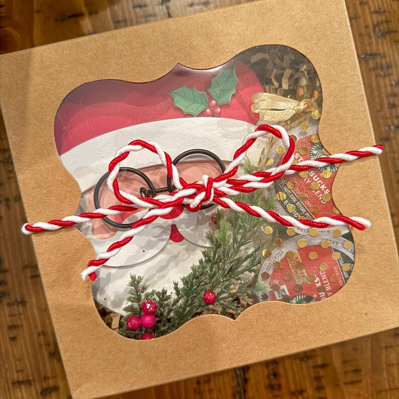 Op de afbeelding: Een bruine kartonnen doos met een transparant venster dat een kerstman-uitsparing, groene takjes, rode bessen en een rode en witte touw strik laat zien. De doos bevat ook een klein pakketje Starbucks-koffie.