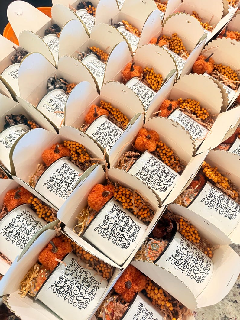 May include: A collection of white gift boxes, each containing a white mug printed with the text Fall leaves, Sweater weather, Hayrides, Bonfire, Pie harvest, S'mores. Each box includes a small orange fabric pumpkin and yellow berry sprigs.