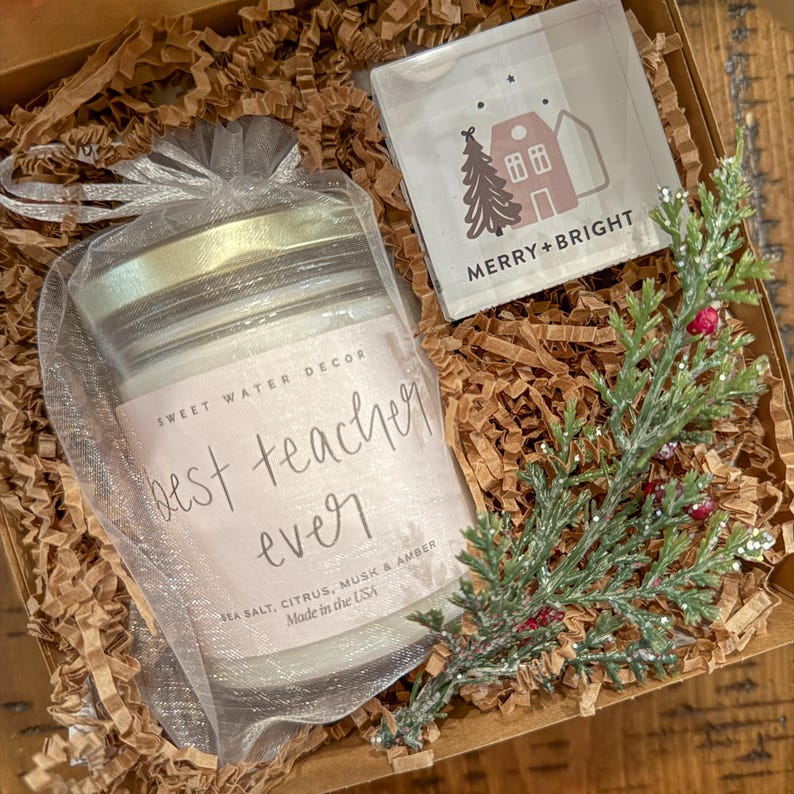 Op de afbeelding: Een cadeaudoos gevuld met bruin kreukpapier, een witte kaars met een roze label met de tekst "best teacher ever", en een klein vierkant glas met een huis- en boomontwerp en de tekst "MERRY + BRIGHT".