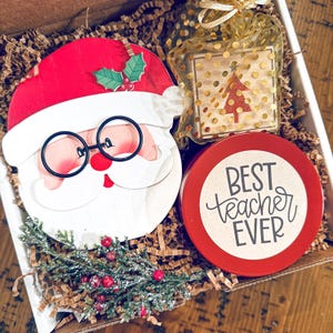 Op de afbeelding: Een cadeaudoos gevuld met bruin kreukpapier, een rood-witte kerstmanversiering, een klein houten blokje met een gouden ster en een rode kerstboom, en een rode kaars met een bruin label waarop "BEST teacher EVER" staat.