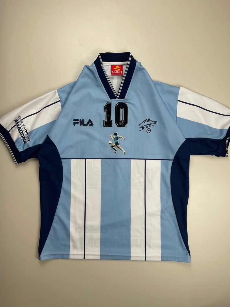 May include: Light blue and white striped soccer jersey with the number 10 on the front. Features the Fila logo, a player graphic, and a signature. The sleeves are white with navy blue trim. The collar is navy blue and white.
