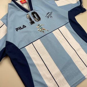 May include: Light blue and white striped soccer jersey with the number 10, a player graphic, and the FILA logo. The jersey has dark blue accents on the sleeves and collar. Includes a signature.