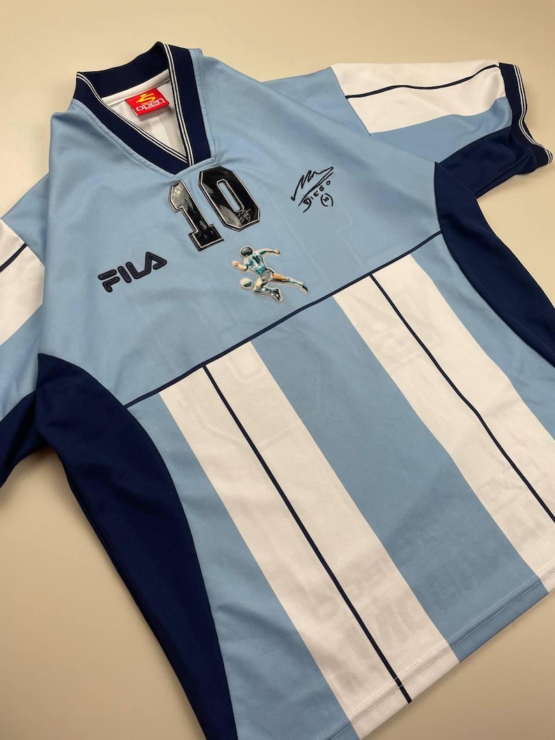 May include: Light blue and white striped soccer jersey with the number 10, a player graphic, and the FILA logo. The jersey has dark blue accents on the sleeves and collar. Includes a signature.