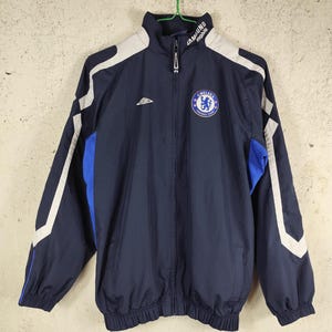 May include: Navy blue zip-up jacket with white and blue panels. The Chelsea Football Club logo is on the chest, and the Samsung Mobile logo is on the collar. It has a full zip and a high collar.