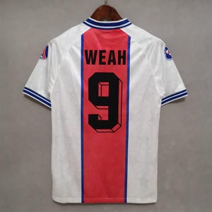 Könnte beinhalten: Ein weißes Fußballtrikot mit einem roten vertikalen Streifen auf der Rückseite, mit dem Namen "WEAH" und der Nummer "9" in Schwarz. Das Trikot hat blaue Verzierungen an Kragen und Ärmeln sowie ein Teamlogo auf den Ärmeln.