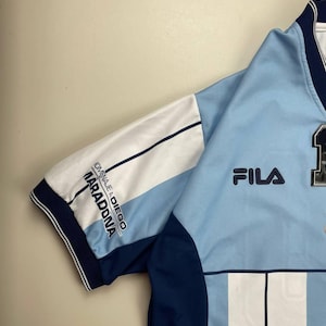 May include: Light blue and white athletic jersey with navy blue accents. The jersey features the Fila logo and the text "HOMENAJE A DIEGO MARADONA" on the sleeve. The jersey has vertical stripes and a number on the front.