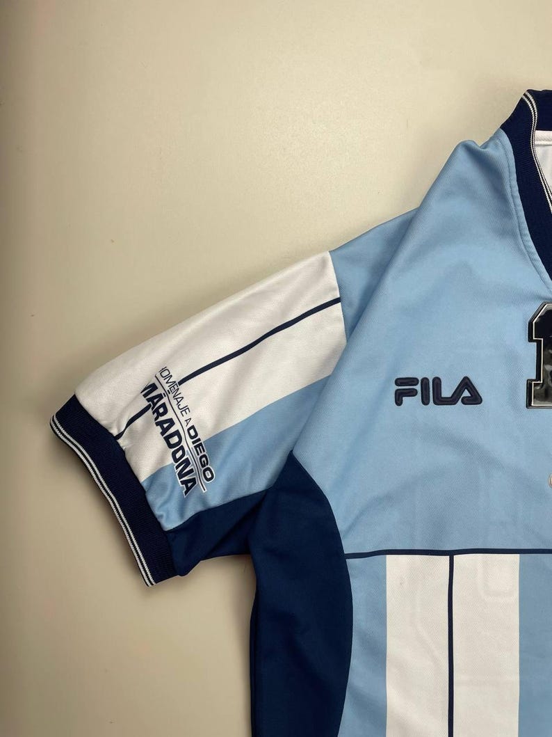 May include: Light blue and white athletic jersey with navy blue accents. The jersey features the Fila logo and the text "HOMENAJE A DIEGO MARADONA" on the sleeve. The jersey has vertical stripes and a number on the front.