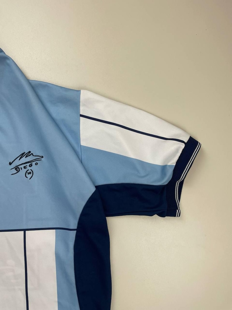 May include: Light blue and white soccer jersey with a navy blue sleeve and trim. The jersey features a signature and the name "DIEGO" with the number "10" printed on the front.
