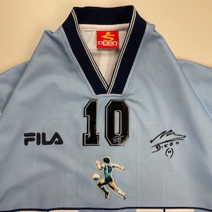 May include: Light blue soccer jersey with the number 10, a player graphic, and the Fila logo. The jersey has a navy blue collar and trim, and the Open Sport logo is visible. The jersey also features a signature.