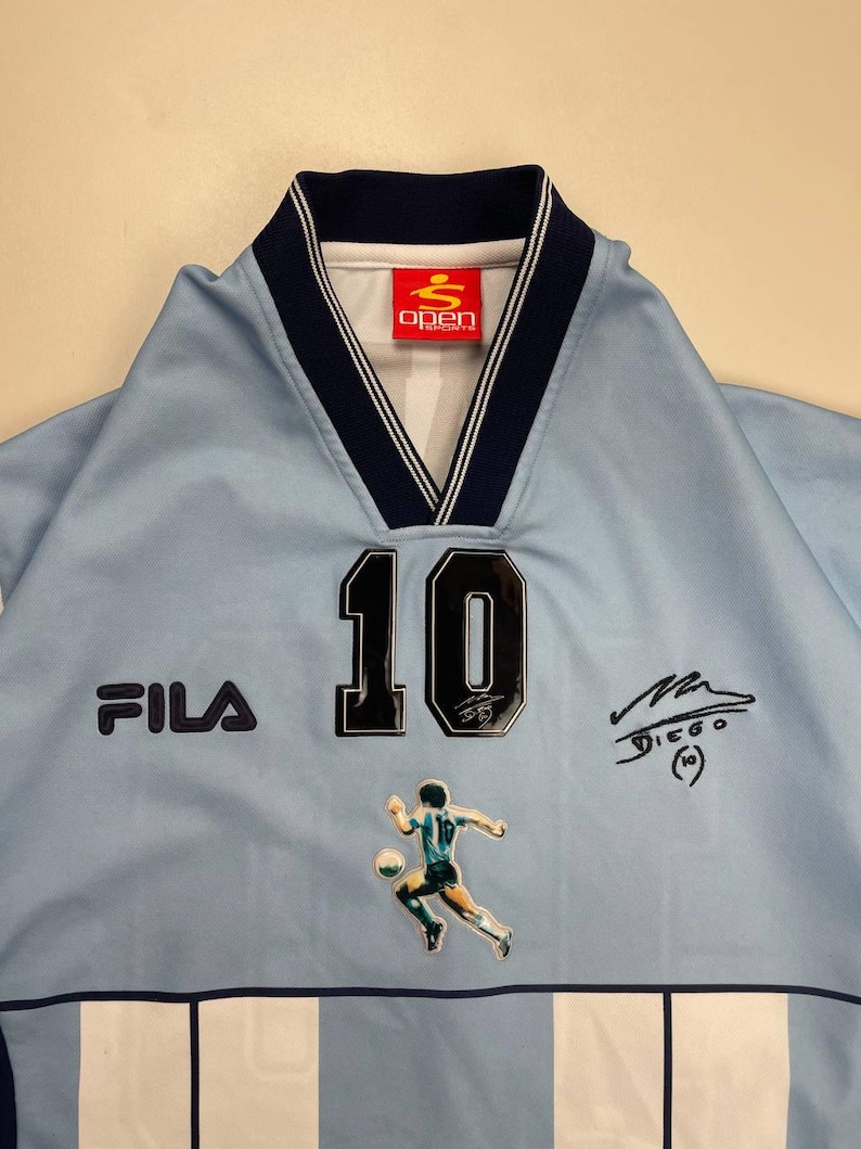 May include: Light blue soccer jersey with the number 10, a player graphic, and the Fila logo. The jersey has a navy blue collar and trim, and the Open Sport logo is visible. The jersey also features a signature.