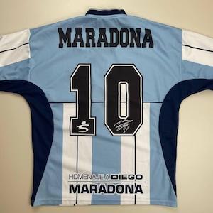 May include: Light blue and white striped soccer jersey with the name "MARADONA" and the number "10" in black. The jersey also features the text "HOMENAJE A DIEGO MARADONA" and a signature.