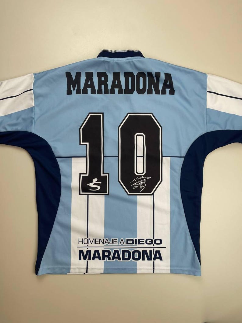 May include: Light blue and white striped soccer jersey with the name "MARADONA" and the number "10" in black. The jersey also features the text "HOMENAJE A DIEGO MARADONA" and a signature.