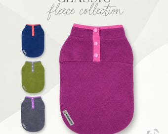 May include: A collection of four sleeveless fleece pet vests in navy blue, olive green, charcoal gray, and magenta. Each vest features a contrasting button placket with three buttons and a small rectangular brand tag labeled MR BOBBETTES.