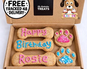 Puede incluir: Una caja de cartón con tres premios para perro en forma de hueso con glaseado rosa y azul que deletrean Happy Birthday Rosie, junto a dos premios en forma de pata decorados con glaseado y virutas. La tapa incluye el texto Mr Bobbettes TREATS y un dibujo.