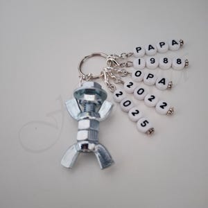 Könnte beinhalten: Ein silberner Schlüsselanhänger mit einer Metallfigur aus Mutter und Schraube. Der Schlüsselanhänger hat mehrere Anhänger mit den Worten "PAPA", "1988", "OPA", "2022" und "2025" darauf.