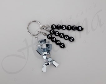 May include: A silver metal keychain with a small figure made of nuts and bolts. The keychain has black beads with white letters spelling "BESTER BONUS PAPA".