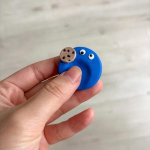 Puede incluir: Un dije circular de arcilla azul con dos ojos móviles blancos con pupilas negras y una pequeña galleta color canela con chispas de chocolate marrón oscuro adherida en la parte superior izquierda de la superficie azul.