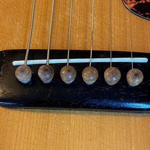 May include: Close-up of a guitar bridge with six tuning pegs. The bridge is made of black wood and has a white plastic strip running across it. The tuning pegs are made of brown wood.