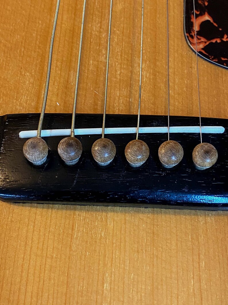 May include: Close-up of a guitar bridge with six tuning pegs. The bridge is made of black wood and has a white plastic strip running across it. The tuning pegs are made of brown wood.