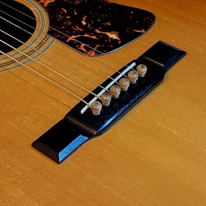 May include: Close-up of an acoustic guitar bridge with six tuning pegs. The bridge is black with a wood grain finish. The tuning pegs are made of wood and have a natural finish.