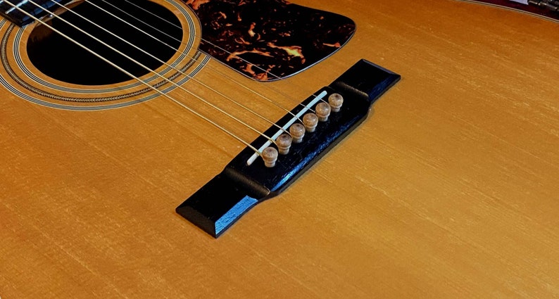 May include: Close-up of an acoustic guitar bridge with six tuning pegs. The bridge is black with a wood grain finish. The tuning pegs are made of wood and have a natural finish.