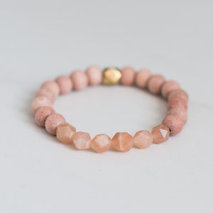 May include: A pink and gold beaded bracelet with a gold accent bead. The bracelet is made of round wooden beads and faceted sunstone beads.