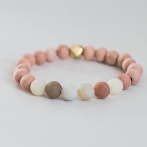 May include: A stretch bracelet composed of round beads in shades of dusty pink, muted brown, and soft cream, featuring a single metallic gold-toned accent bead. The beads are arranged in a circular pattern, creating a continuous loop of earthy tones.