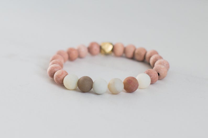 May include: A stretch bracelet composed of round beads in shades of dusty pink, muted brown, and soft cream, featuring a single metallic gold-toned accent bead. The beads are arranged in a circular pattern, creating a continuous loop of earthy tones.