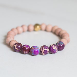 May include: A stretch bracelet featuring a series of matte, light pink, round beads interspersed with five polished, deep purple, marbled stone beads and a single, small, metallic gold-toned bead centered at the top of the circular arrangement.
