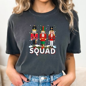May include: Dark gray t-shirt featuring three colorful nutcracker figures with the word "SQUAD" in white and red. The design includes snowflakes and festive details, perfect for holiday wear.