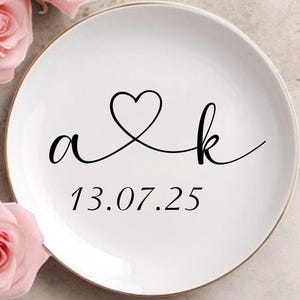 May include: A white ceramic plate with a gold rim features the letters "a & k" connected by a heart, and the date "13.07.25" in black script. Three pink roses with green leaves are arranged around the plate.