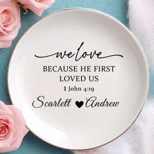 May include: A white ceramic plate with a gold rim, featuring the text "we love BECAUSE HE FIRST LOVED US 1 John 4:19 Scarlett ♥ Andrew". The plate is surrounded by pink roses and rests on a blue surface.