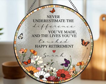 Op de afbeelding: Een ronde glazen zonnevanger met een zwart metalen frame en een ophangketting. In het midden staat de tekst 'NEVER UNDERESTIMATE THE DIFFERENCE YOU'VE MADE, AND THE LIVES YOU'VE TOUCHED HAPPY RETIREMENT' met de naam 'Sarah' en een rood hartje.