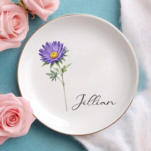 K&ouml;nnte beinhalten: Wei&szlig;e Keramikplatte mit goldenem Rand, mit einer lila Blumenillustration und dem Namen "Jillian" in Schreibschrift. Rosa Rosen und gr&uuml;ne Bl&auml;tter sind um die Platte auf einer blauen Oberfl&auml;che angeordnet.