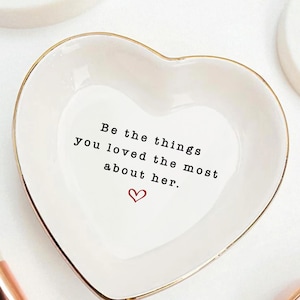 May include: White heart-shaped trinket dish with gold trim. The dish has the text "Be the things you loved the most about her." in black, with a small red heart below the text. Makeup brushes and other cosmetic items are in the background.