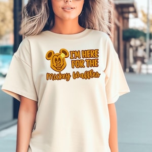 May include: Cream-colored t-shirt featuring a Mickey Mouse waffle design with the text "I'M HERE FOR THE Mickey Waffles" in brown and yellow. The shirt has short sleeves and a relaxed fit.