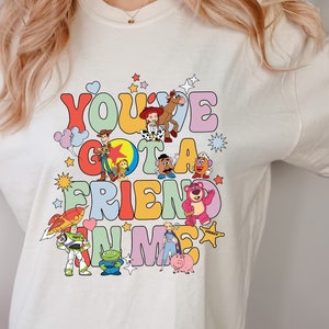 May include: Cream-colored t-shirt featuring a colorful graphic of characters from the movie "Toy Story." The text "You've Got a Friend in Me" is displayed in a retro font. The design includes Woody, Buzz Lightyear, and other characters.