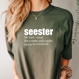 May include: A green t-shirt with the word "seester" defined as "like a sister, only cooler." The text is in white and the shirt has short sleeves.