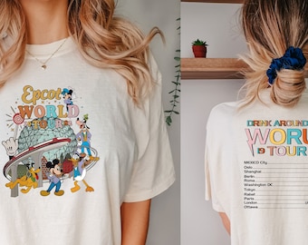 Puede incluir: Una camiseta blanca con un gráfico de Mickey Mouse y otros personajes de Disney en la parte delantera. La parte posterior de la camiseta dice "Drink Around the World Tour" con una lista de países y ciudades.